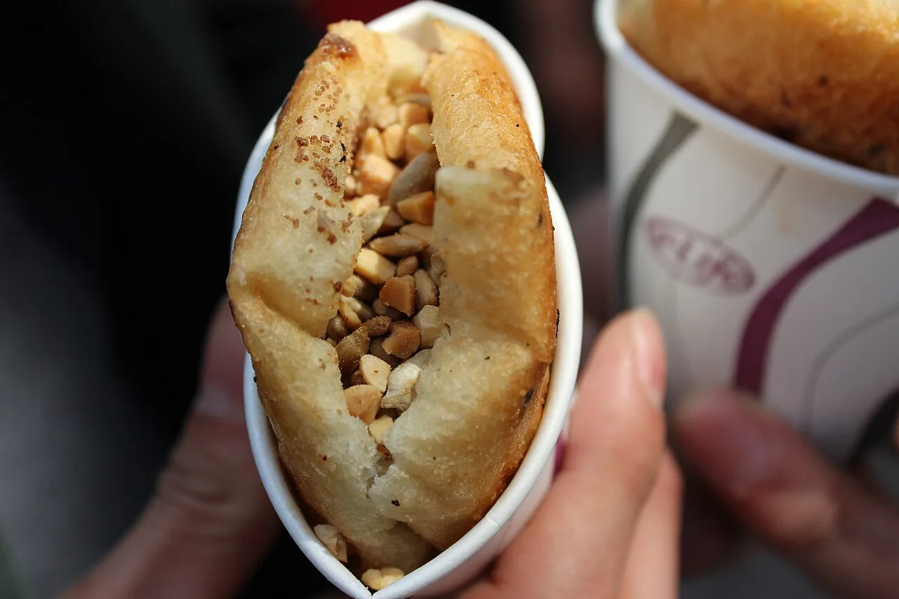 A hand holding ssiat hotteok, a fried pancake split open and stuffed with seeds and nuts, in a paper cup