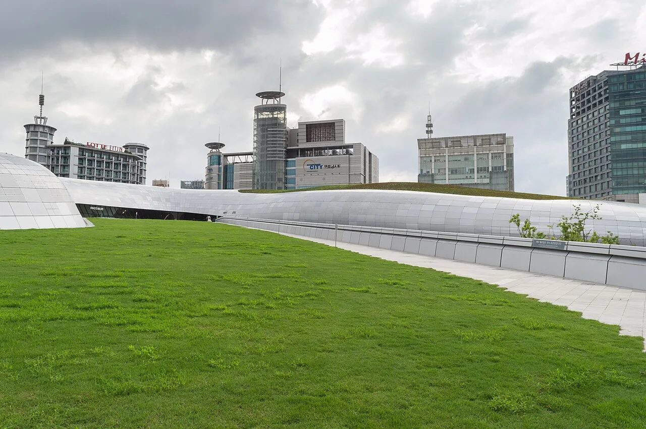 The grassy rooftop of Dongdaemun Design Plaza flowing down to the plaza with city towers behind