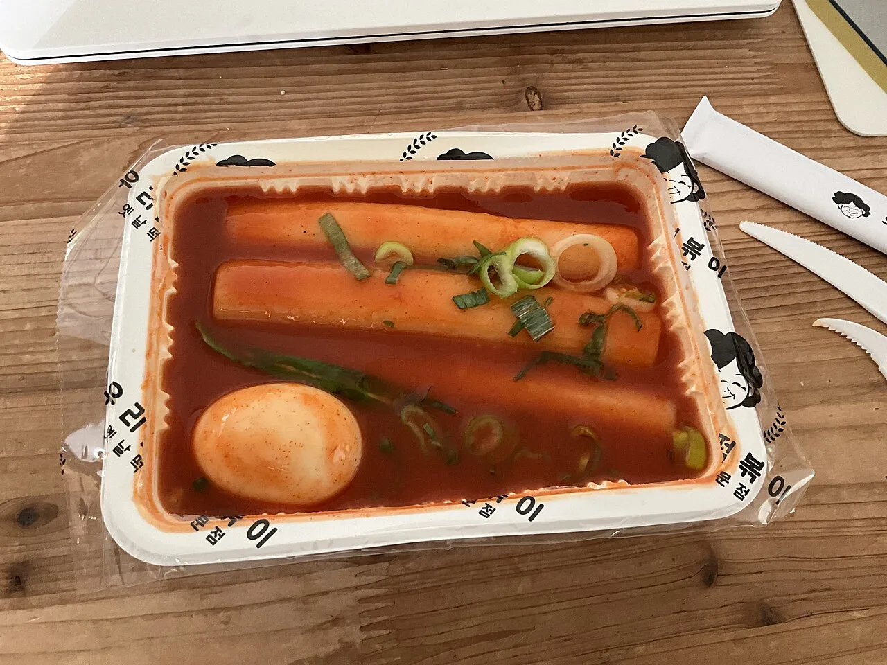 A takeaway tray of tteokbokki rice cakes in red sauce with a boiled egg and fish cake